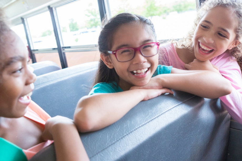 Three students sit on a bus leaning over a seat. All three are smiling.