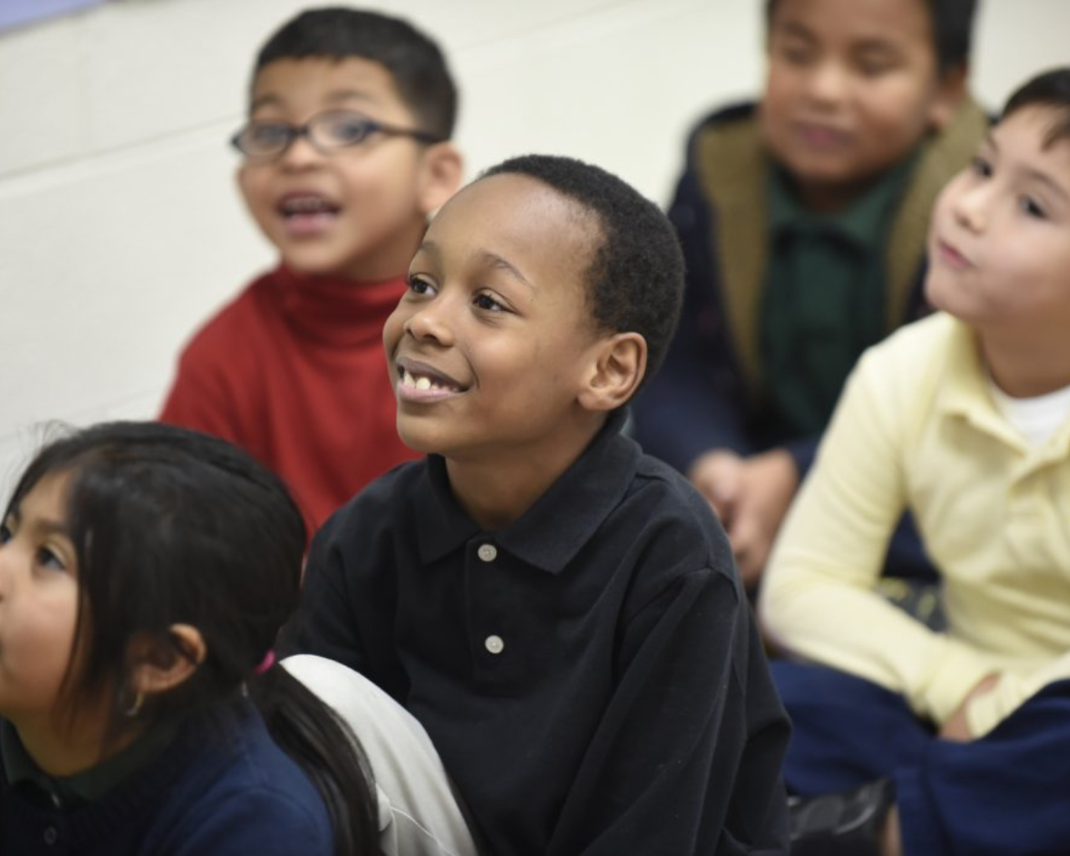 Multiple students sit on a classroom rug listening to a teacher (not pictured). One student in the middle has a smile and bright, hopeful eyes.